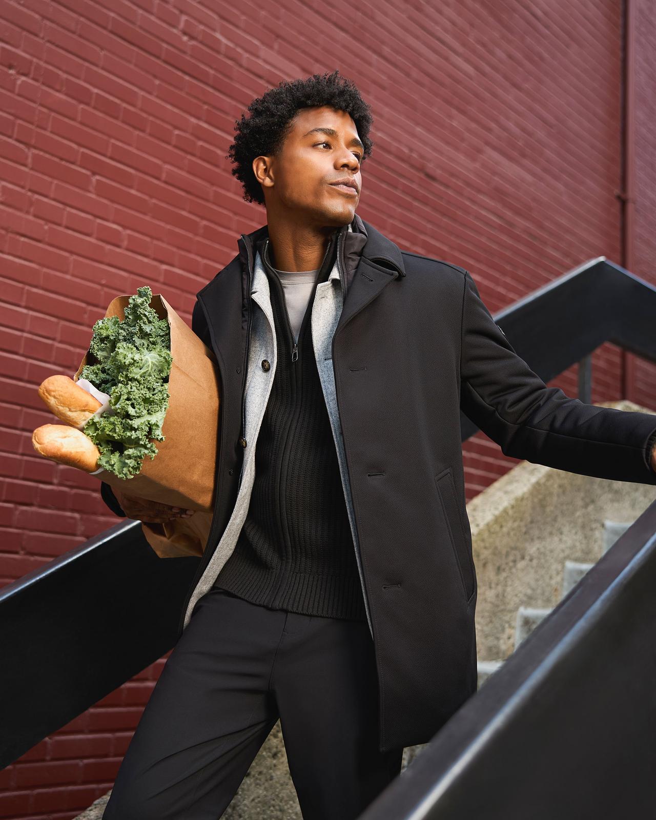 Man wearing a black coat layered over a black sweater and pants, holding a grocery bag