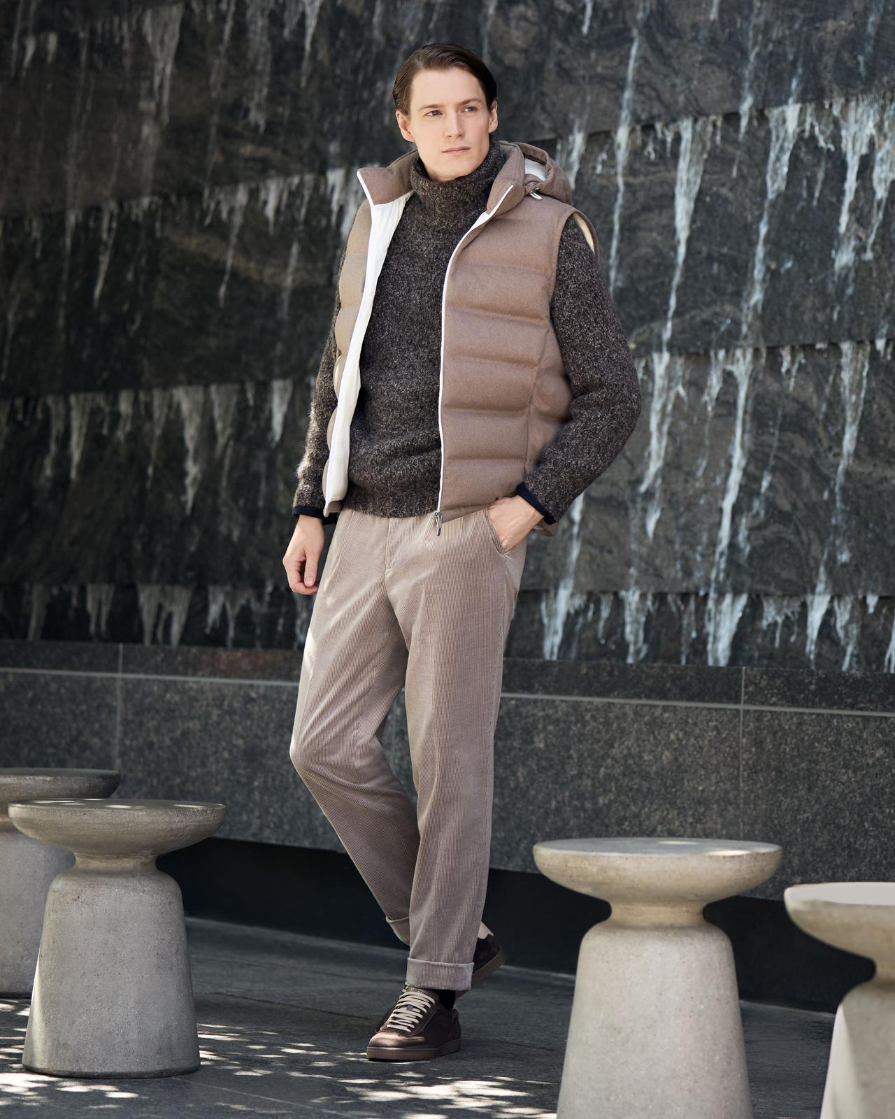 Homme portant un gilet matelassé brun, un chandail foncé et un pantalon beige, debout devant un mur de pierre