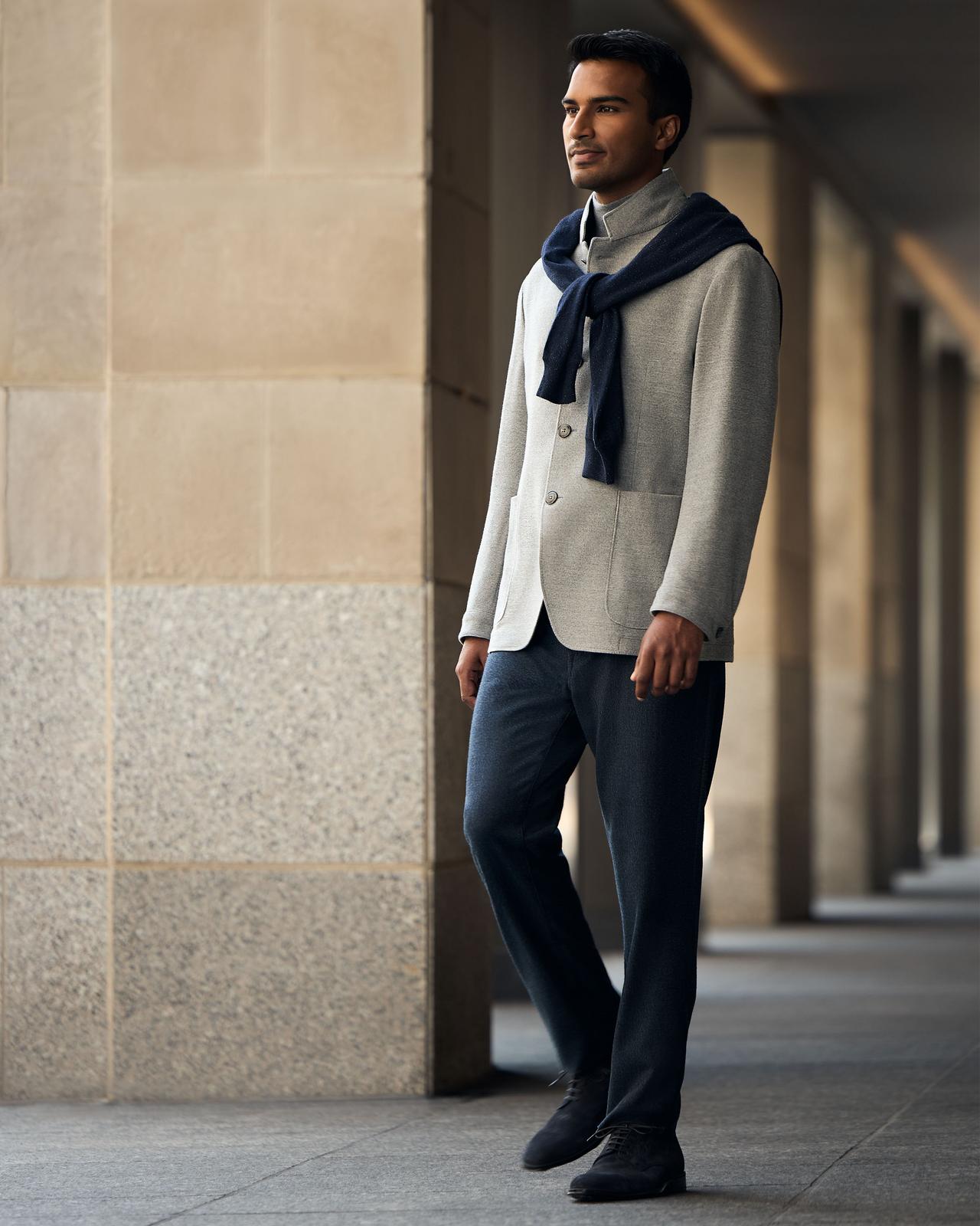 A man wearing a light grey jacket with a navy sweater draped over his shoulders and dark trousers