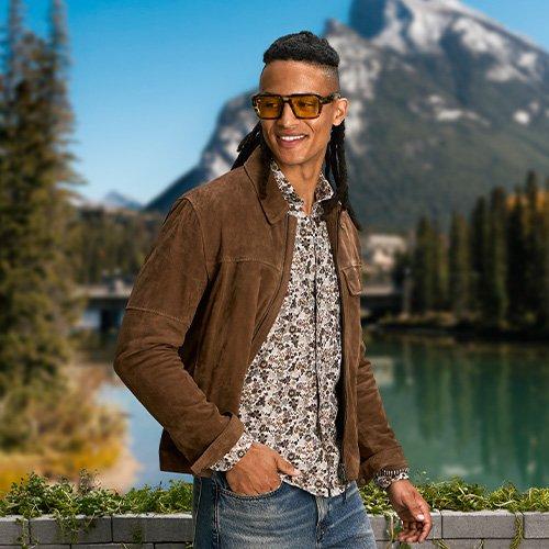 male model wearing sunglasses, dress shirt, jeans and overshirt outside infront of mountain scenery