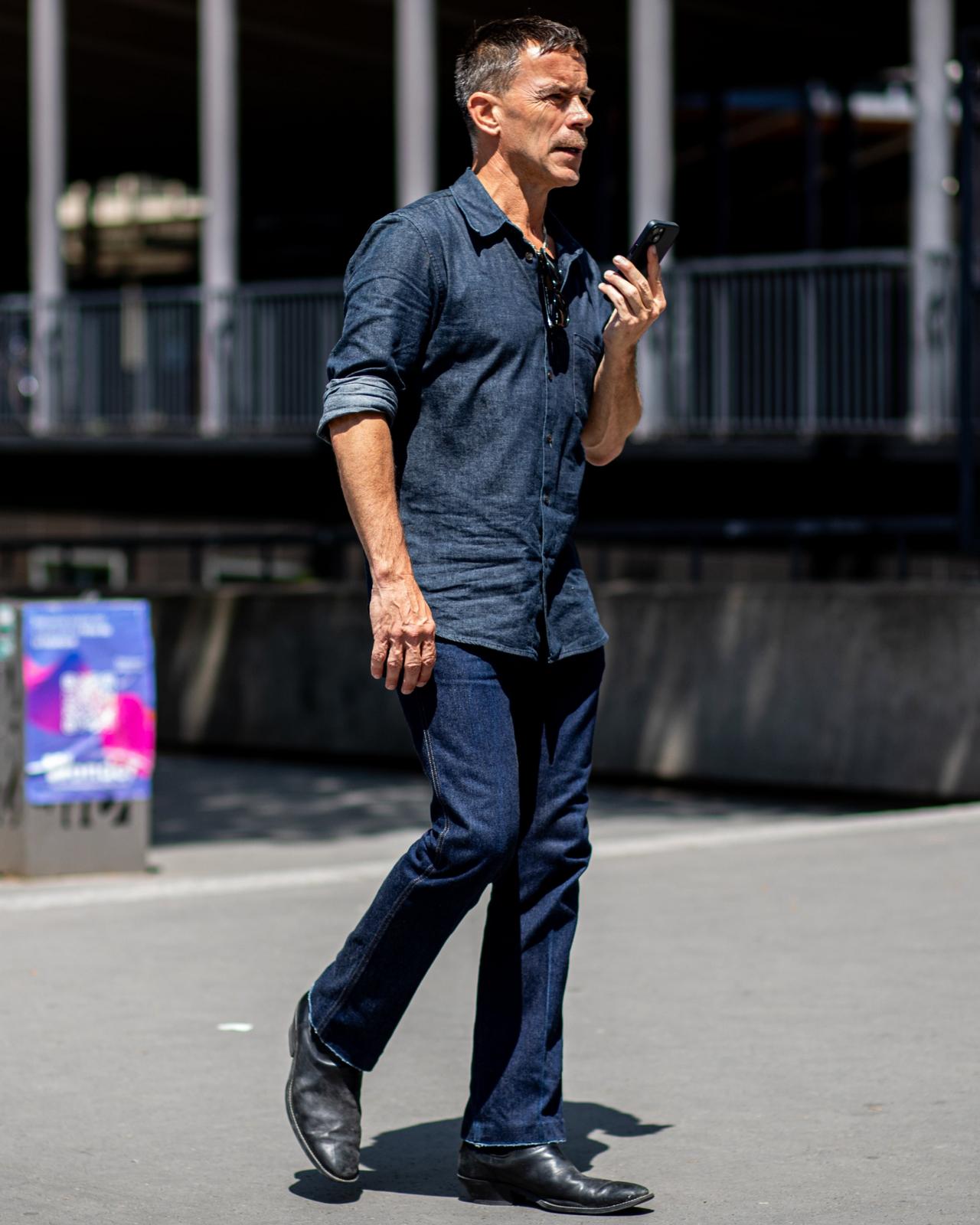 A man wearing a dark denim shirt with rolled-up sleeves, paired with dark jeans and black leather shoes, walking outdoors