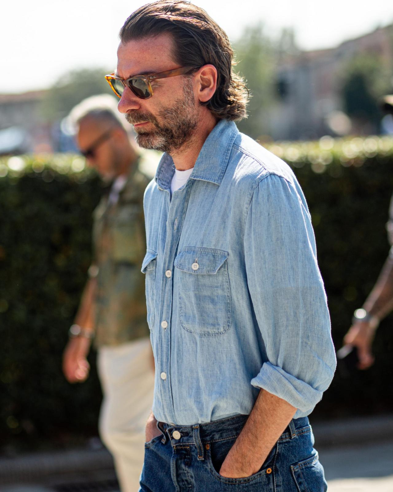 A man wearing a light denim shirt with rolled-up sleeves, paired with dark jeans
