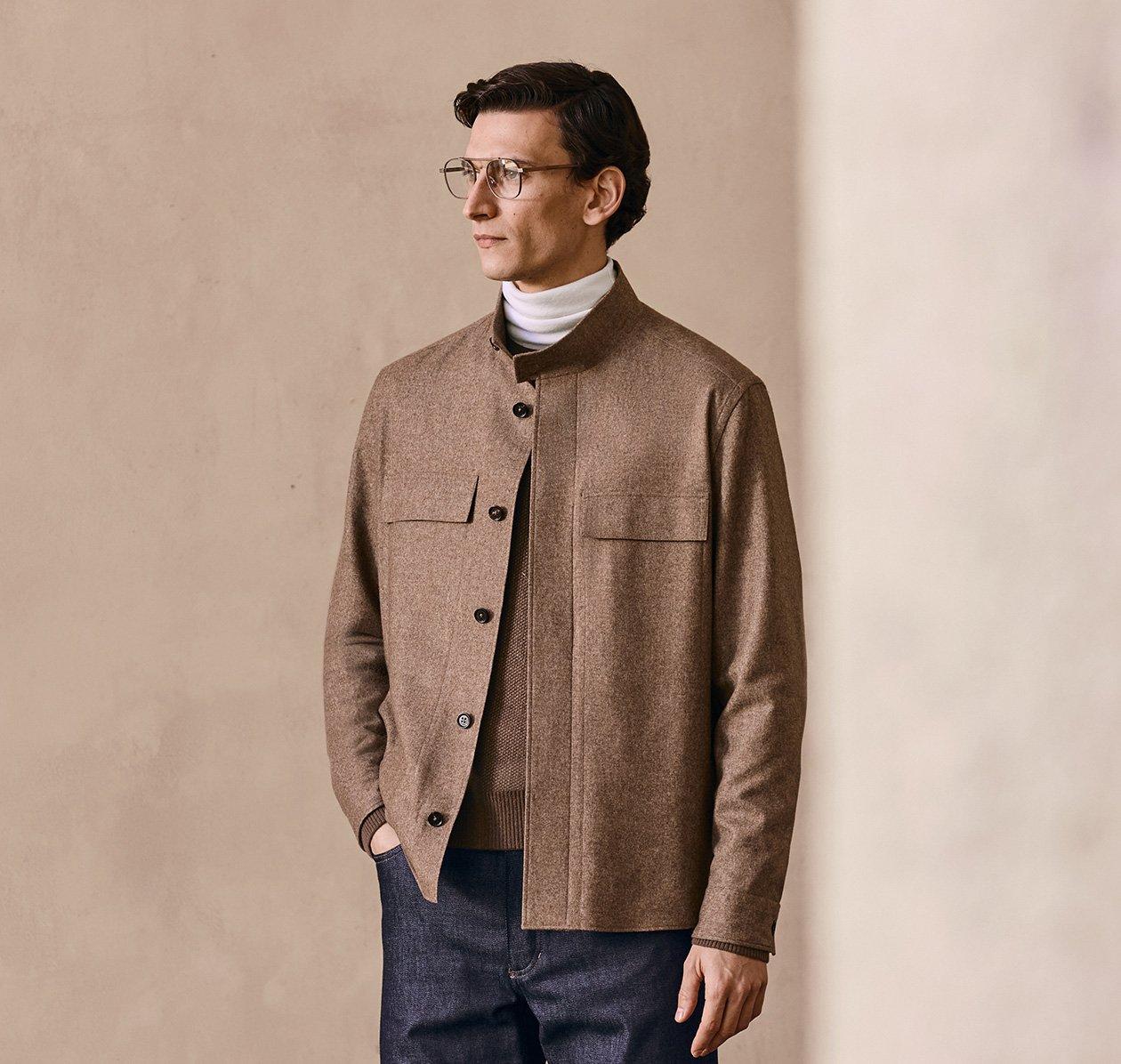 A man wearing a beige button-up jacket over a white turtleneck and dark trousers