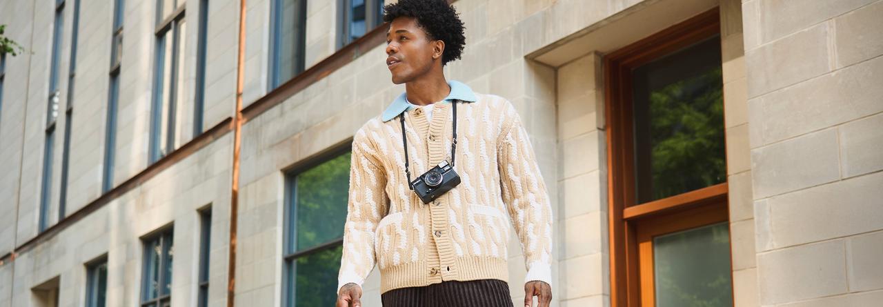 Homme portant un cardigan beige à motifs avec un appareil photo au cou, debout à l’extérieur