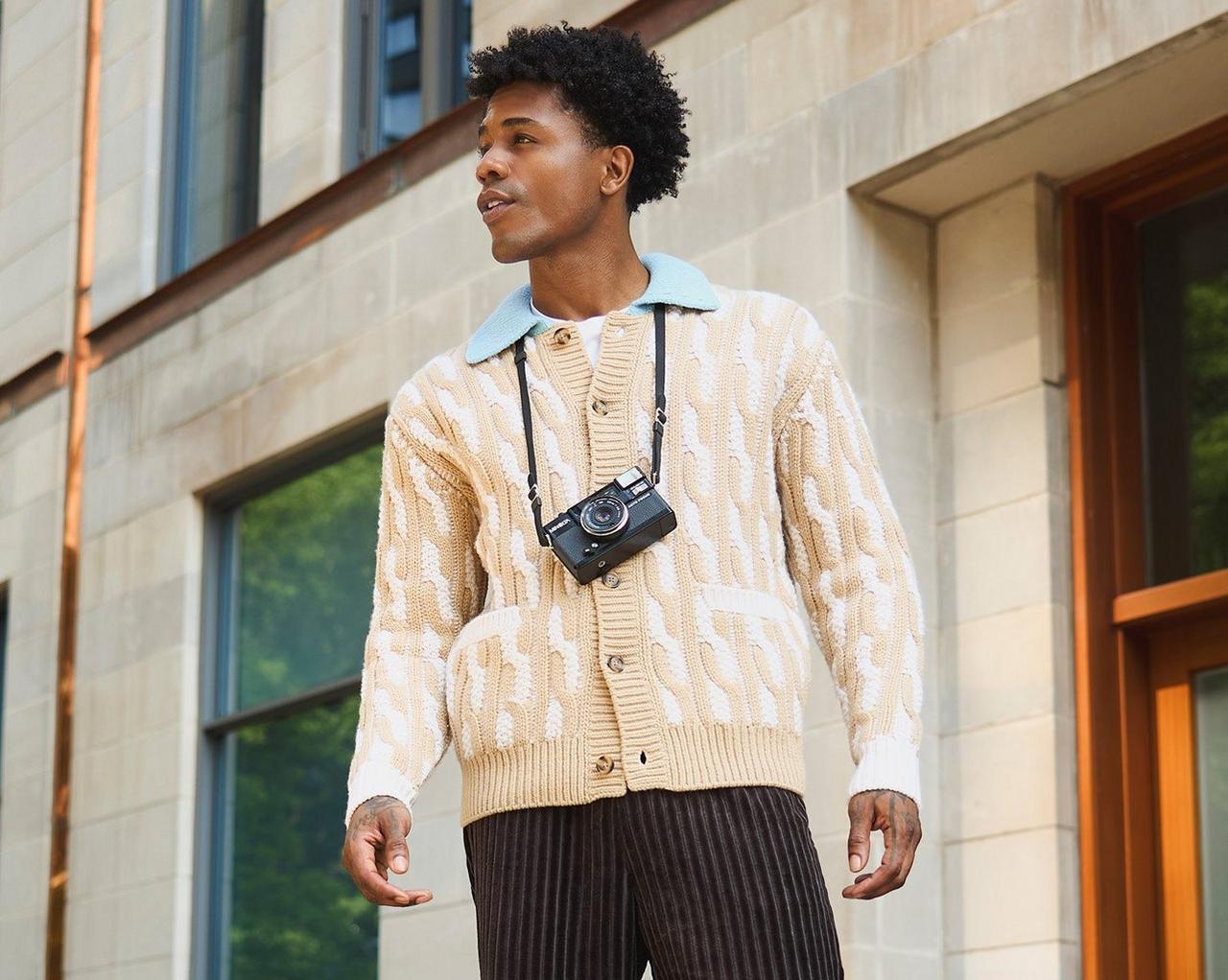 Homme portant un cardigan beige à motifs avec un appareil photo au cou, debout à l’extérieur