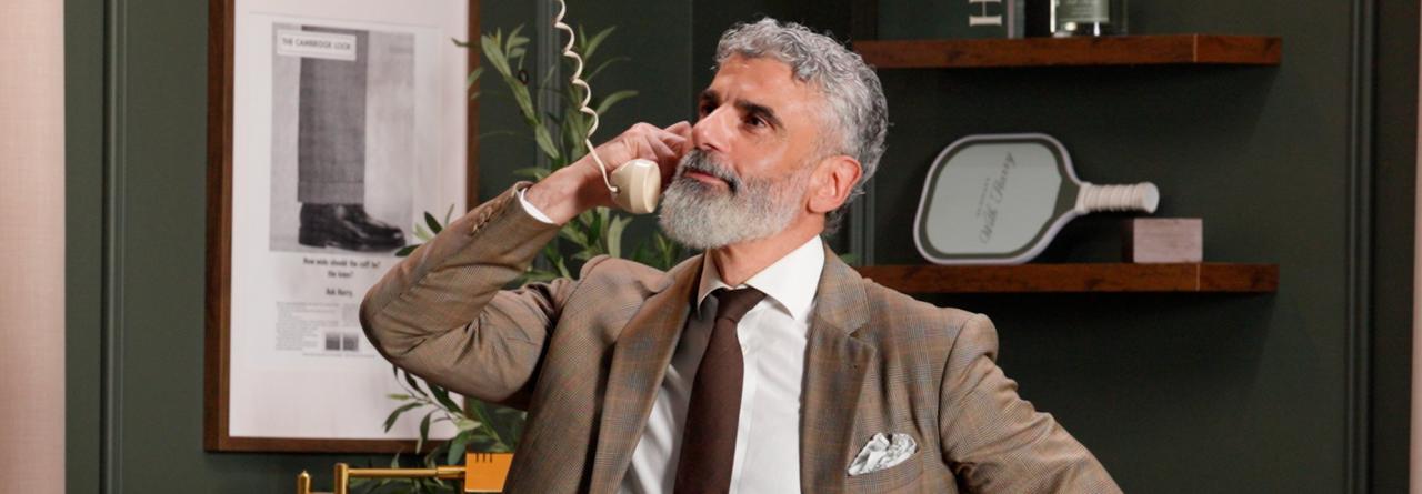 A man in a brown plaid suit and tie speaking on a vintage rotary phone in a modern office setting
