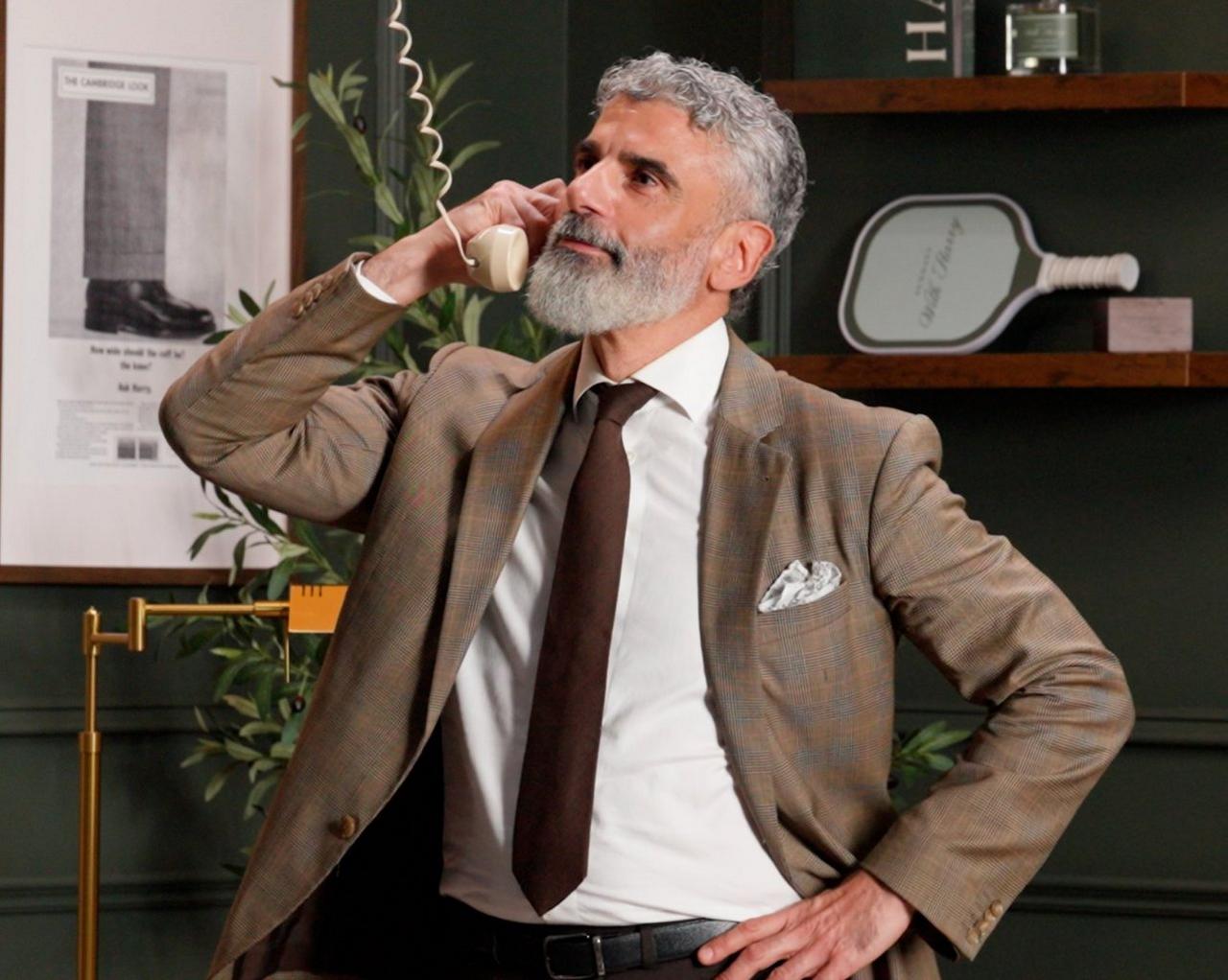 Un homme en costume à carreaux bruns et cravate, parlant au téléphone à cadran rotatif vintage dans un bureau moderne.