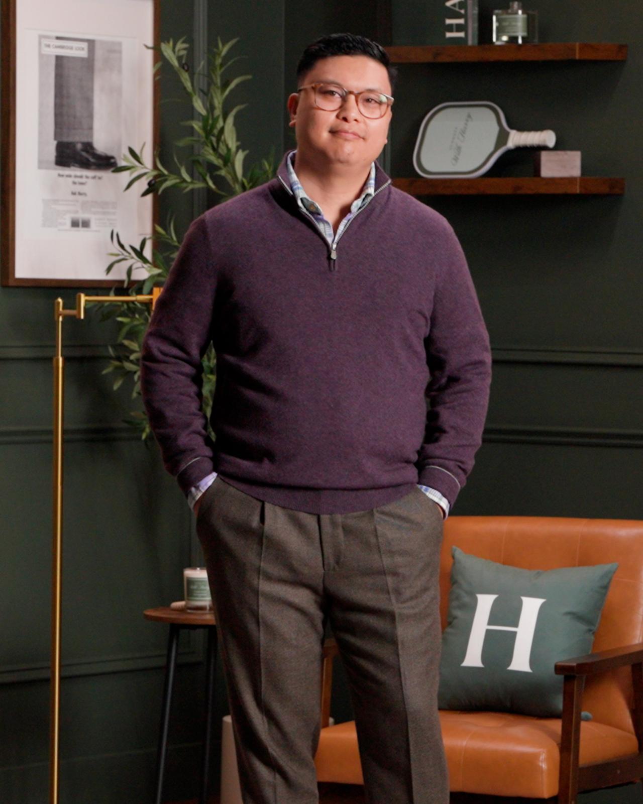 A man standing indoors wearing a deep purple quarter-zip sweater layered over a collared shirt, paired with tailored gray trousers