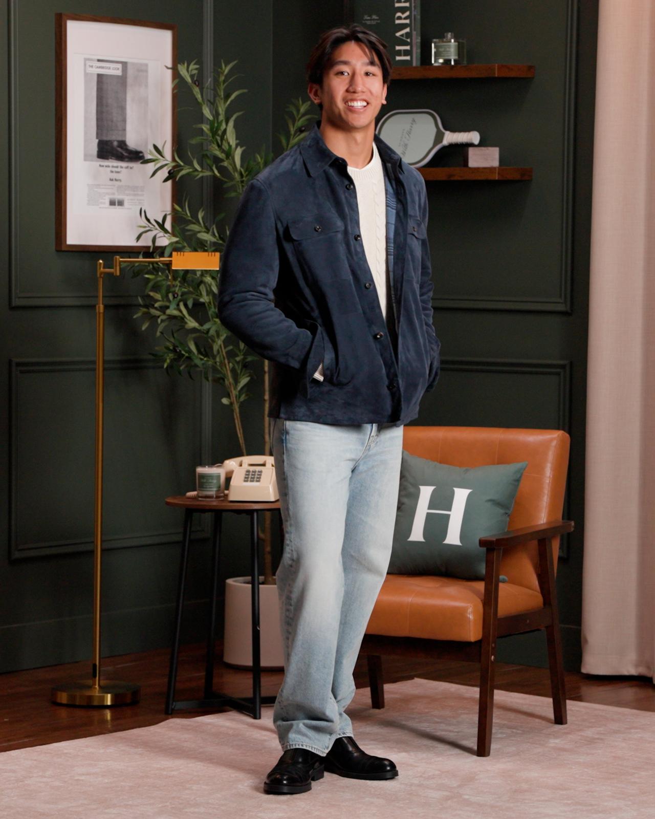 A man standing in a stylish living room, wearing a navy suede jacket over a white shirt paired with light blue jeans and black dress shoes