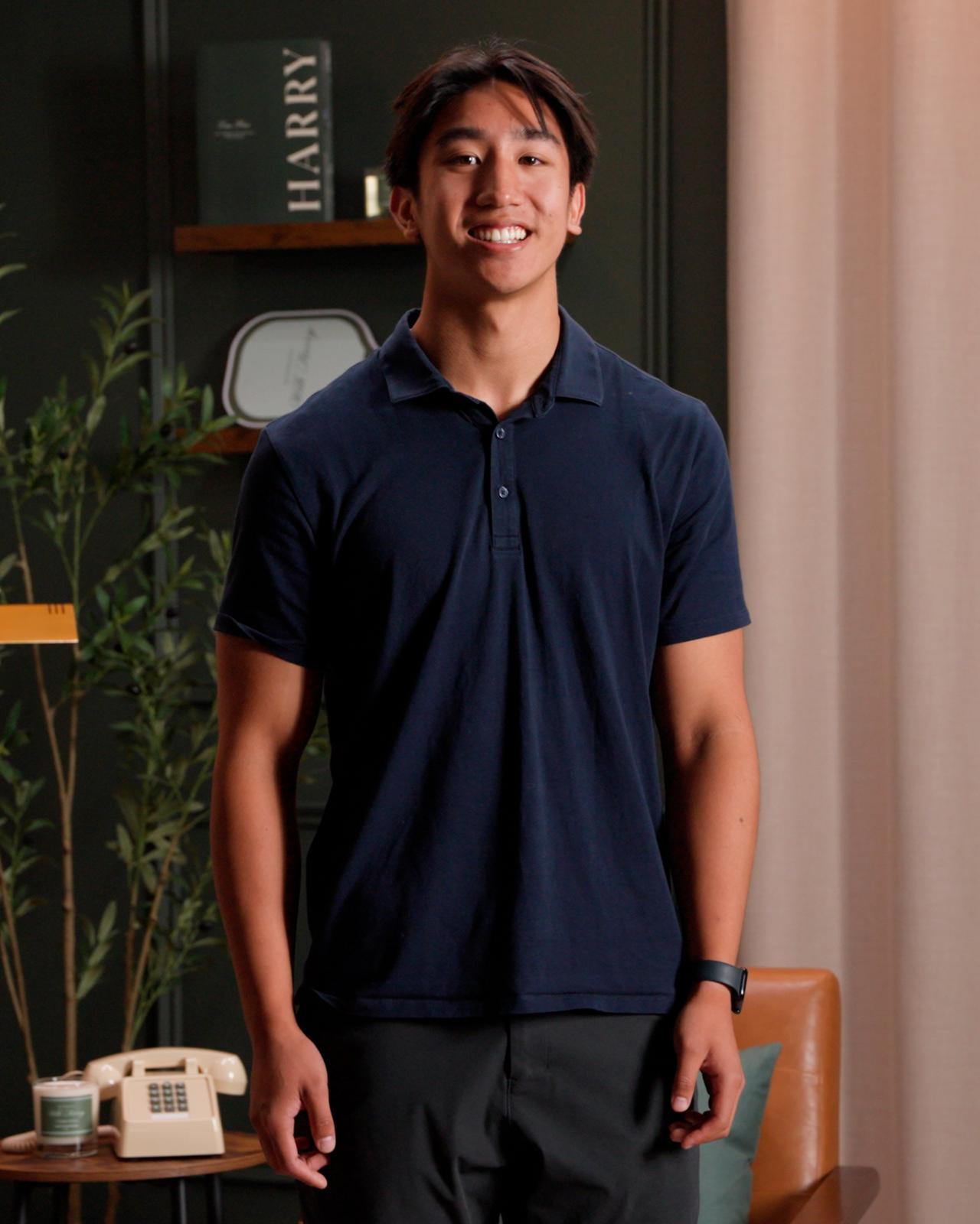 A man wearing a navy short-sleeve polo shirt paired with dark trousers, standing indoors near a small round table with a vintage-style telephone