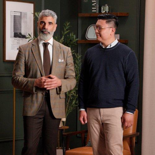 Deux hommes se tenant debout dans une pièce élégamment décorée, l’un vêtu d’un blazer à carreaux bruns avec une pochette blanche, et l’autre portant un chandail bleu marine et un pantalon beige