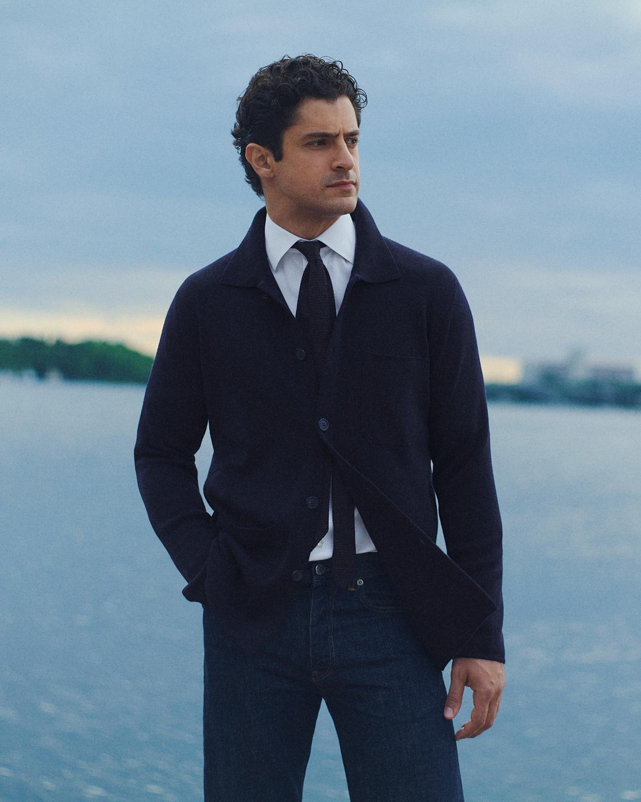 Man wearing a white dress shirt and dark knit tie under a navy knit overshirt with dark jeans, standing by a lake, Eton Week, FW26