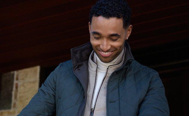 Homme souriant appuyé sur une rampe à l'extérieur, portant une veste matelassée vert foncé sur un col roulé crème, microsite LW Outerwear v2, collection A/H26
