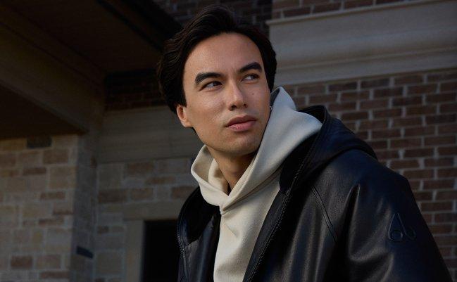 Man wearing a black leather jacket over a cream hoodie, leaning against a black car outdoors, LW Outerwear, FW26 collection