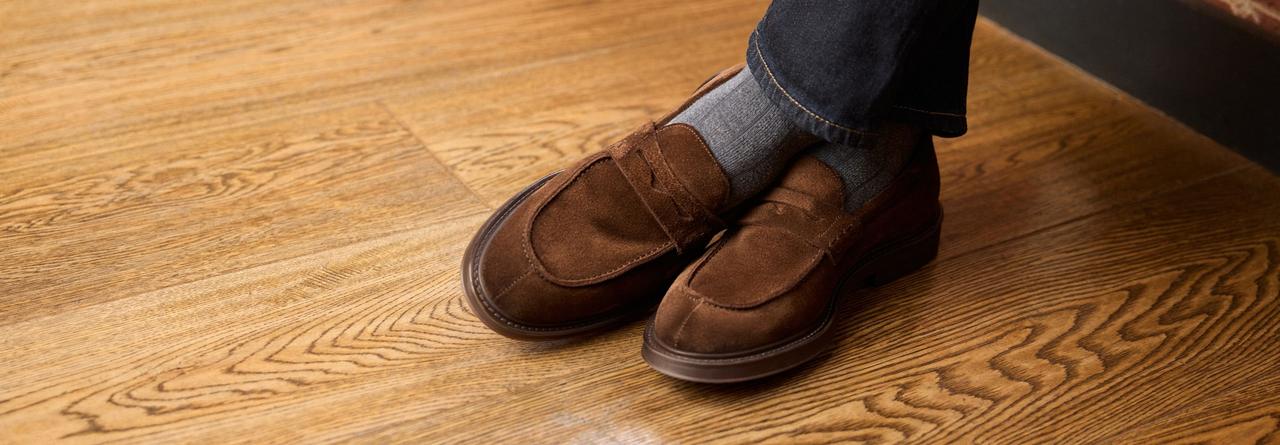 Une paire de mocassins en suède brun portés avec des chaussettes grises et un jean foncé, debout sur un plancher en bois poli