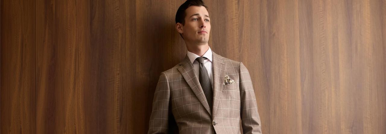 Un homme portant un blazer beige à carreaux bien ajusté, accompagné d’une chemise habillée blanche, d’une cravate foncée et d’un mouchoir de poche décoratif