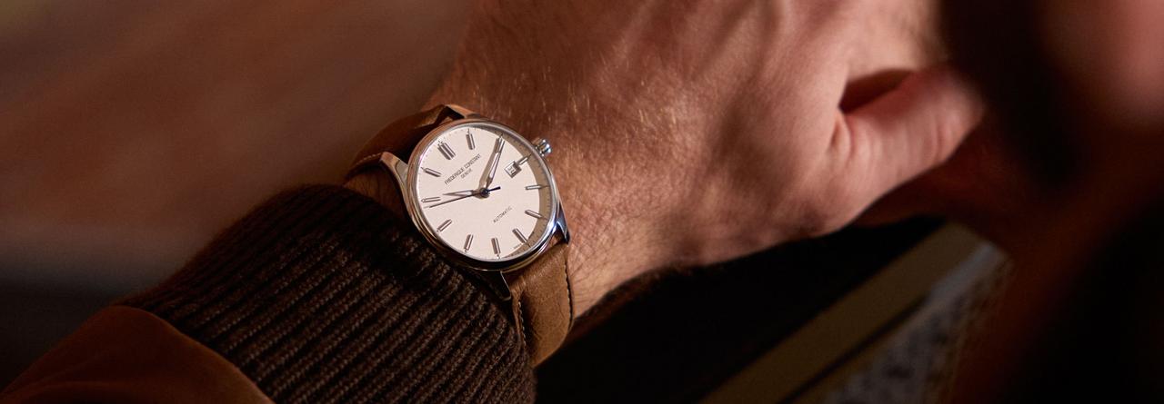 A wrist adorned with an analog wristwatch featuring a white dial, Roman numeral hour markers, and a brown leather strap