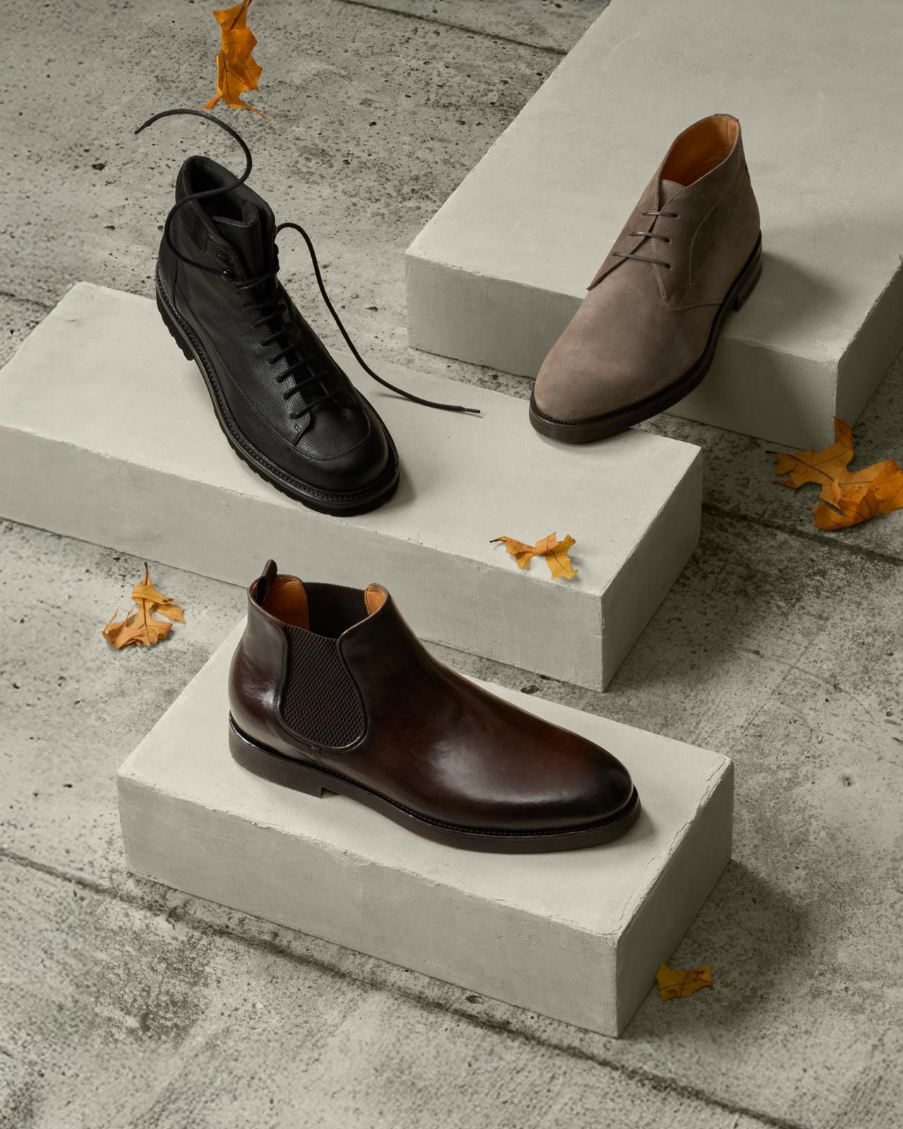 Trois bottes pour homme — à lacets noire, chukka en suède taupe et Chelsea bordeaux — présentées sur des marches en béton, Boots, collection Chaussures A/H26