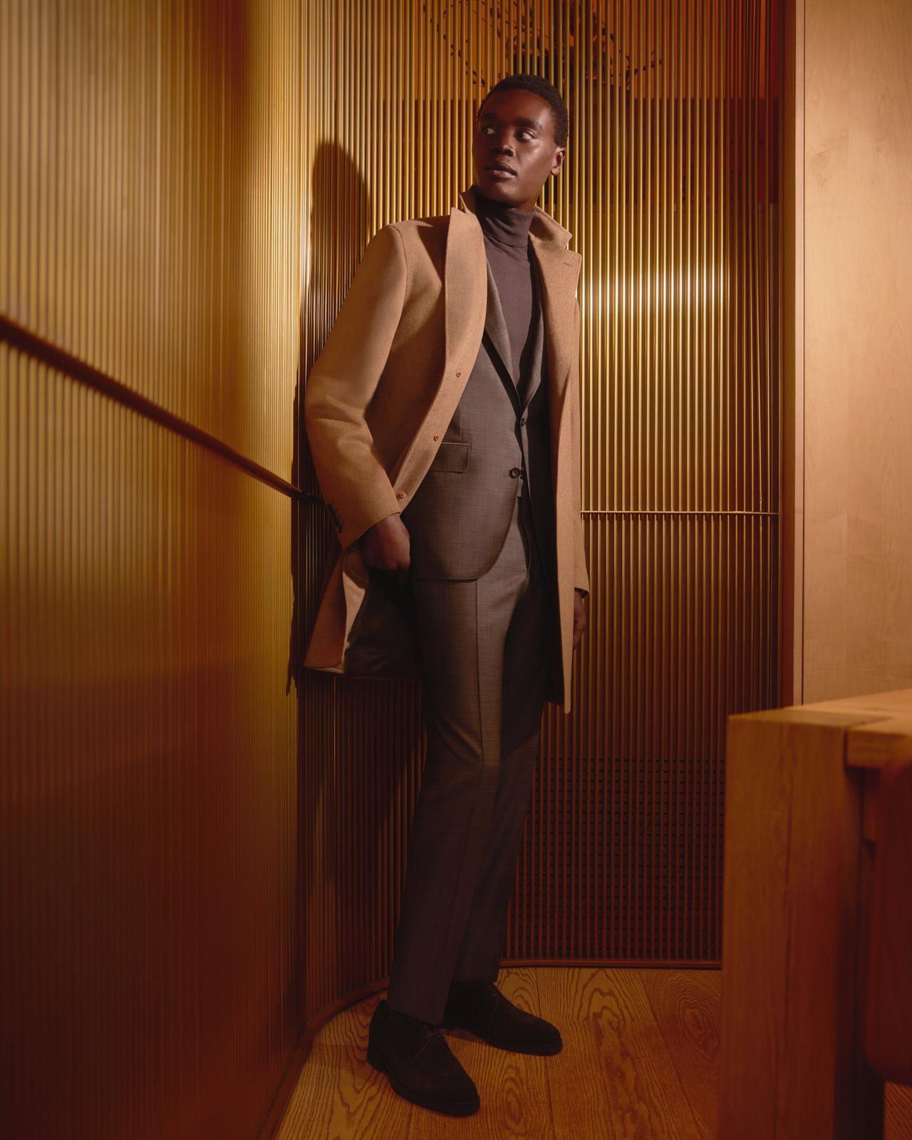  A man in a tailored grey suit paired with a camel-colored overcoat, standing in a warmly lit wooden interior