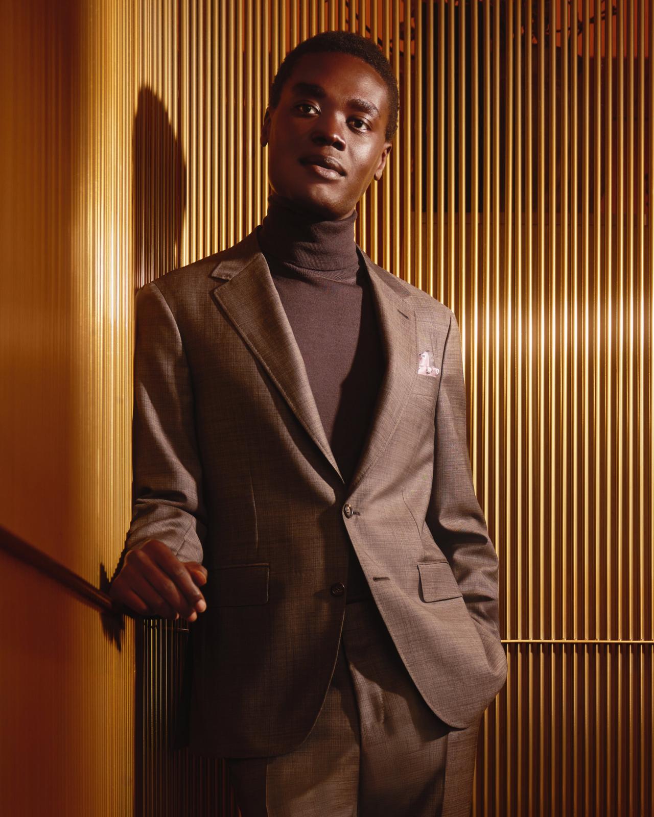 A man wearing a tailored brown suit paired with a dark turtleneck, standing against a backdrop of vertical wooden slats