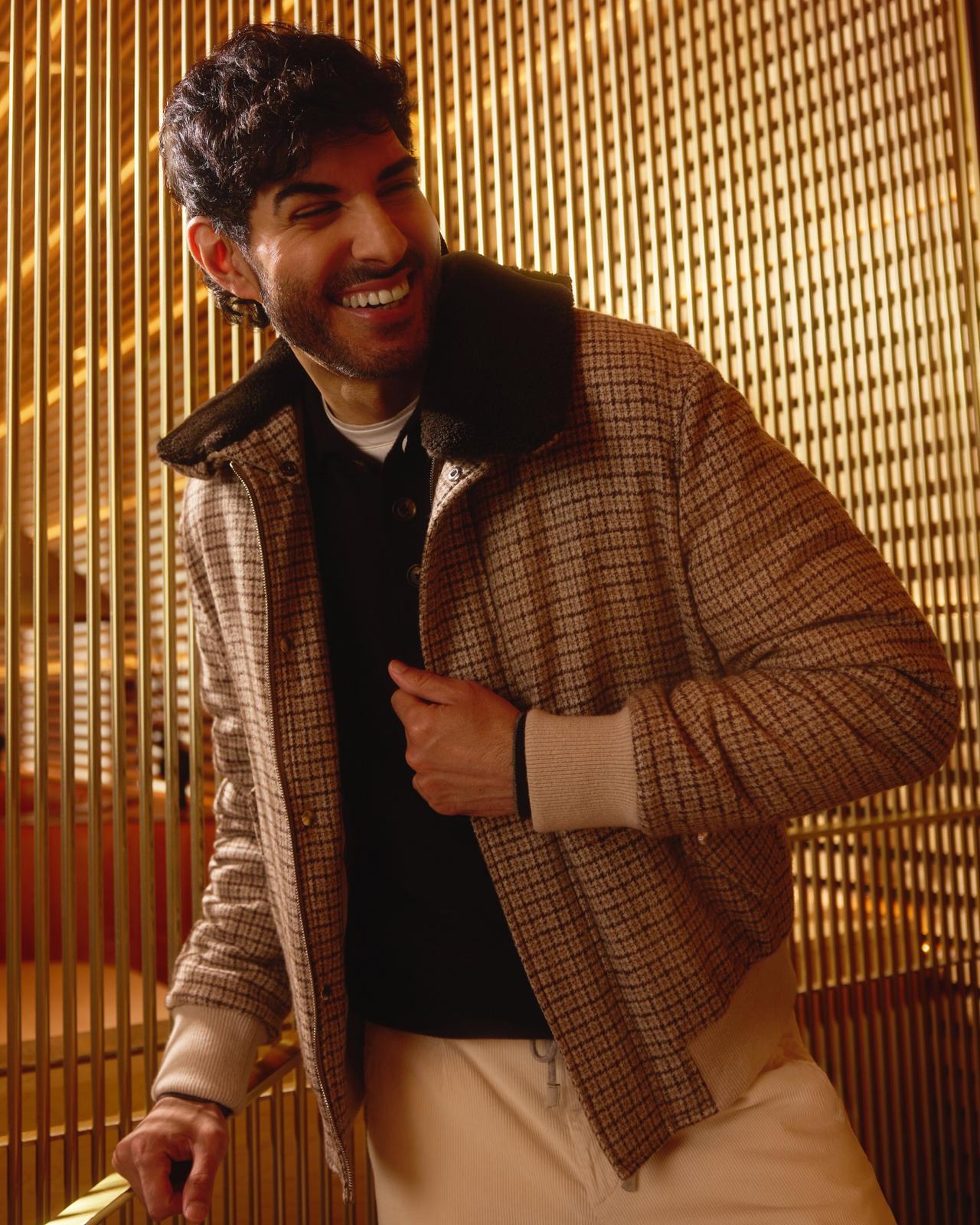 A man wearing a brown plaid bomber jacket with a shearling collar, paired with a black shirt and cream trousers