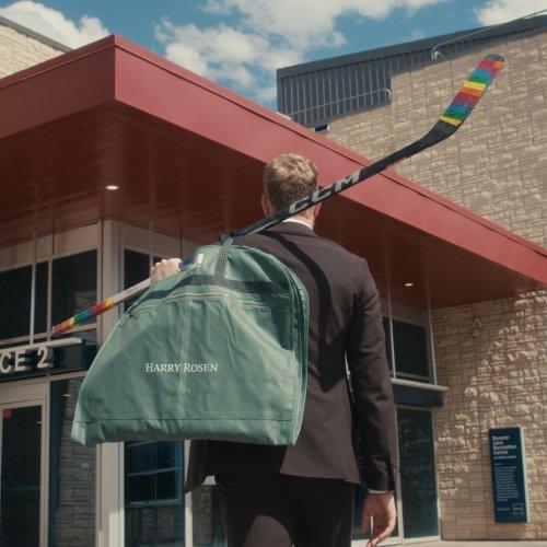 Luke Prokop carrying Harry Rosen labeled green suit bag and and a hockey stick over his shoulder