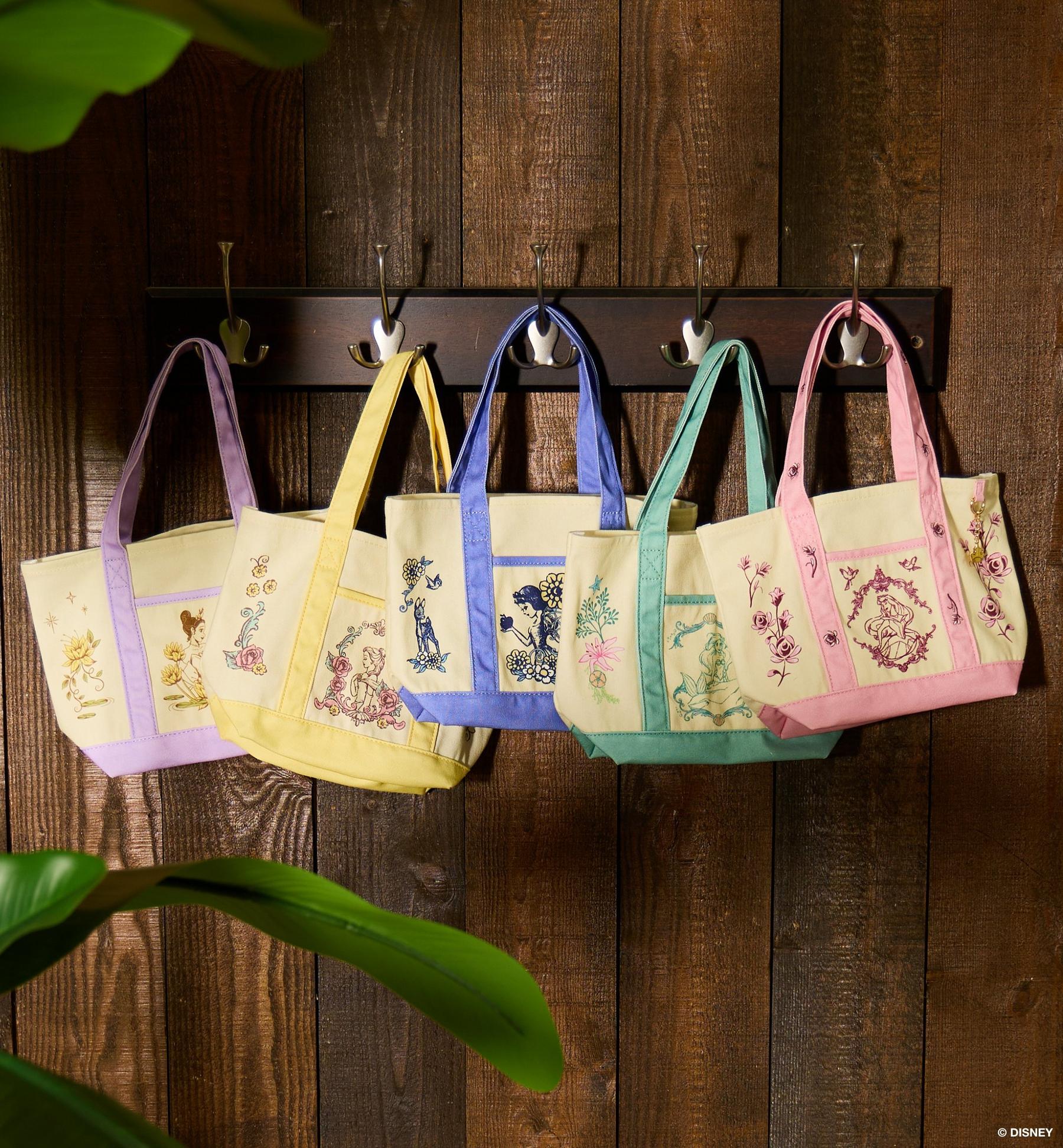 Five Disney illustrated tote bags with pastel handles hanging on wall hooks. Shop bags.