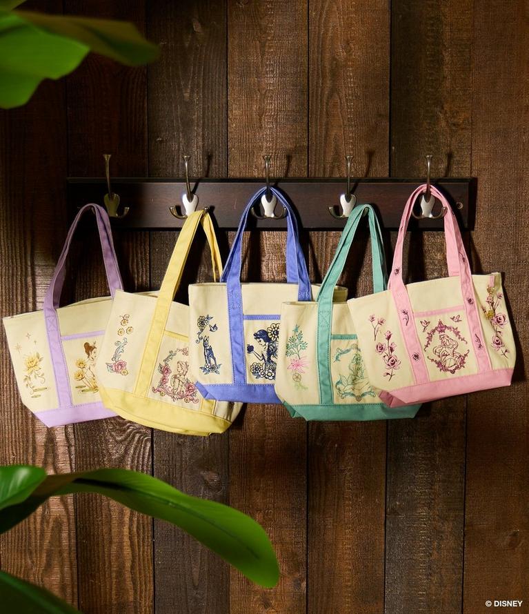 Five embroidered tote bags with pastel handles hanging on wall hooks. Shop bags.