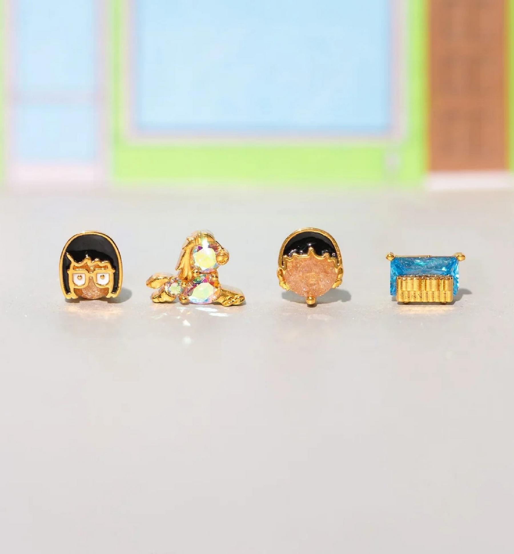 Four gold-tone stud earrings: cartoon face, unicorn, face silhouette, and blue rectangular gem. Shop jewelry.