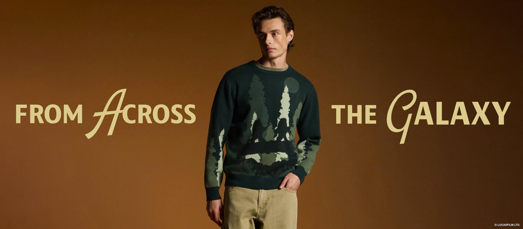 Male model wearing a forest graphic sweater and tan pants with “From Across the Galaxy” text. Shop Star Wars.