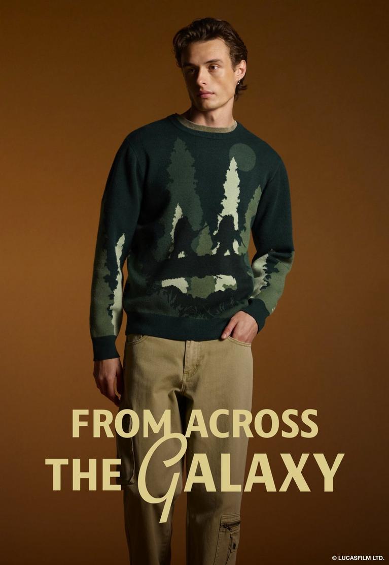 Male model wearing a dark green forest graphic sweater and khaki pants, with “FROM ACROSS THE GALAXY” text. Shop Star Wars.