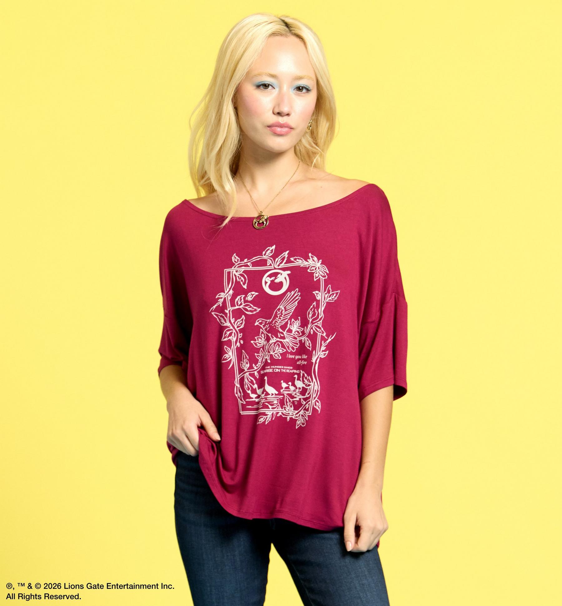 Model wearing a burgundy off-shoulder graphic T-shirt with bird and leaf print. Shop The Hunger Games.