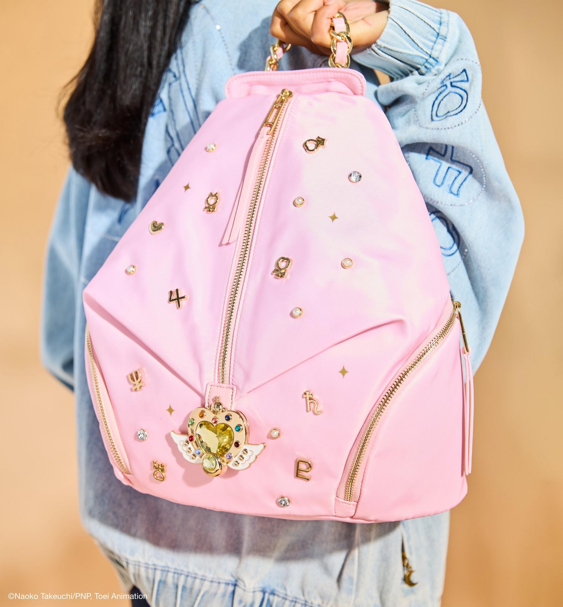 Pink backpack with gold zippers, charms and crystal studs, held by a person in a denim jacket. Shop bags.
