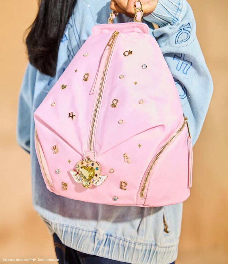 Person holding a pink backpack with gold zippers, charms, and a heart wing emblem. Shop bags.
