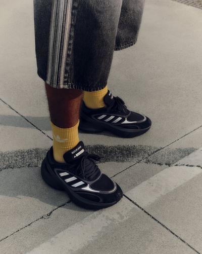 Close-up of black Adidas Goukana sneakers with silver stripes, paired with yellow Adidas socks and grey denim shorts. The shot is taken from a high angle on a concrete pavement.