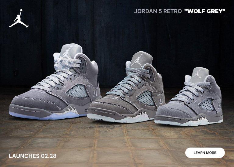 An advertisement for Jordan 5 Retro Wolf Grey sneakers featuring three different sizes of the grey suede shoes displayed in a row. The background is a dark, industrial floor and wall. Text includes the Jordan logo, the shoe name, LAUNCHES 02.28, and a LEARN MORE button.