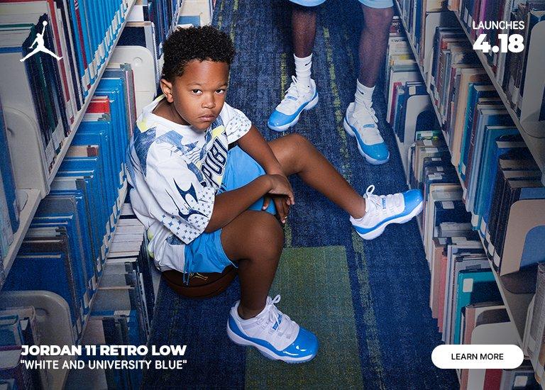 Promotional banner for the Jordan 11 Retro Low "White and University Blue" featuring a child sitting in a library aisle wearing the sneakers. A close-up on the left highlights the white leather upper and vibrant blue patent leather mudguard. Text indicates the shoe "Launches 4.18" with a "Learn More" button in the corner.