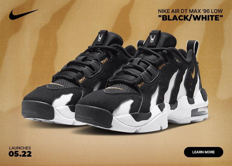 A mobile marketing banner features a pair of Nike Air DT Max 96 Low Black/White sneakers against a textured, light tan and brown background. The athletic shoes showcase a bold, flame-like black and white torn-paper design with gold accents. A black Nike swoosh sits in the top-left corner, while text in the top-right displays the shoe model. The bottom corners include the launch date 05.22 and a black learn more button.