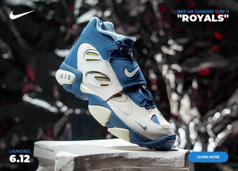 A Nike promotional banner features a single Nike Air Diamond Turf II Royals sneaker balanced on a white stone block against a crumpled, metallic silver and black background with hints of red reflection. The mid-top sneaker showcases a white and deep blue upper with a midfoot strap featuring a white swoosh, side cutouts, and an AIR logo on the heel midsole. A white Nike swoosh is in the top left corner, blue text in the bottom left reads LAUNCHES 6.12, and text in the top right reads NIKE AIR DIAMOND TURF II ROYALS. A blue LEARN MORE button sits in the bottom right corner.