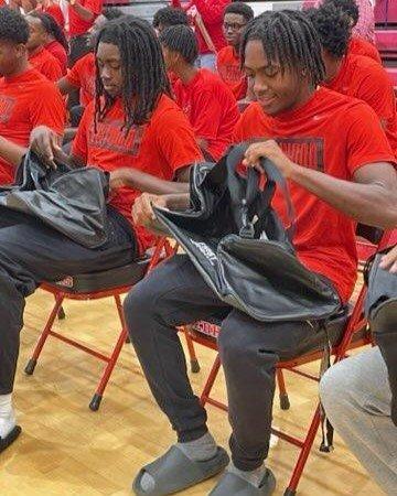 Male student-athletes wearing red shirts seated in the gym, actively unzipping and inspecting their new black Nike duffel bags.
