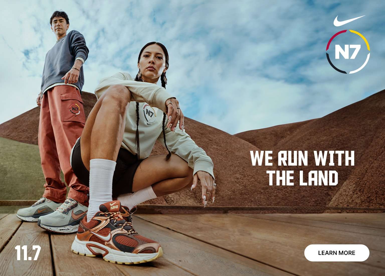 Nike N7 campaign image featuring two people in athletic wear and new shoes on a wooden surface with rolling brown hills in the background. Text reads: 'N7, WE RUN WITH THE LAND, 11.7.'