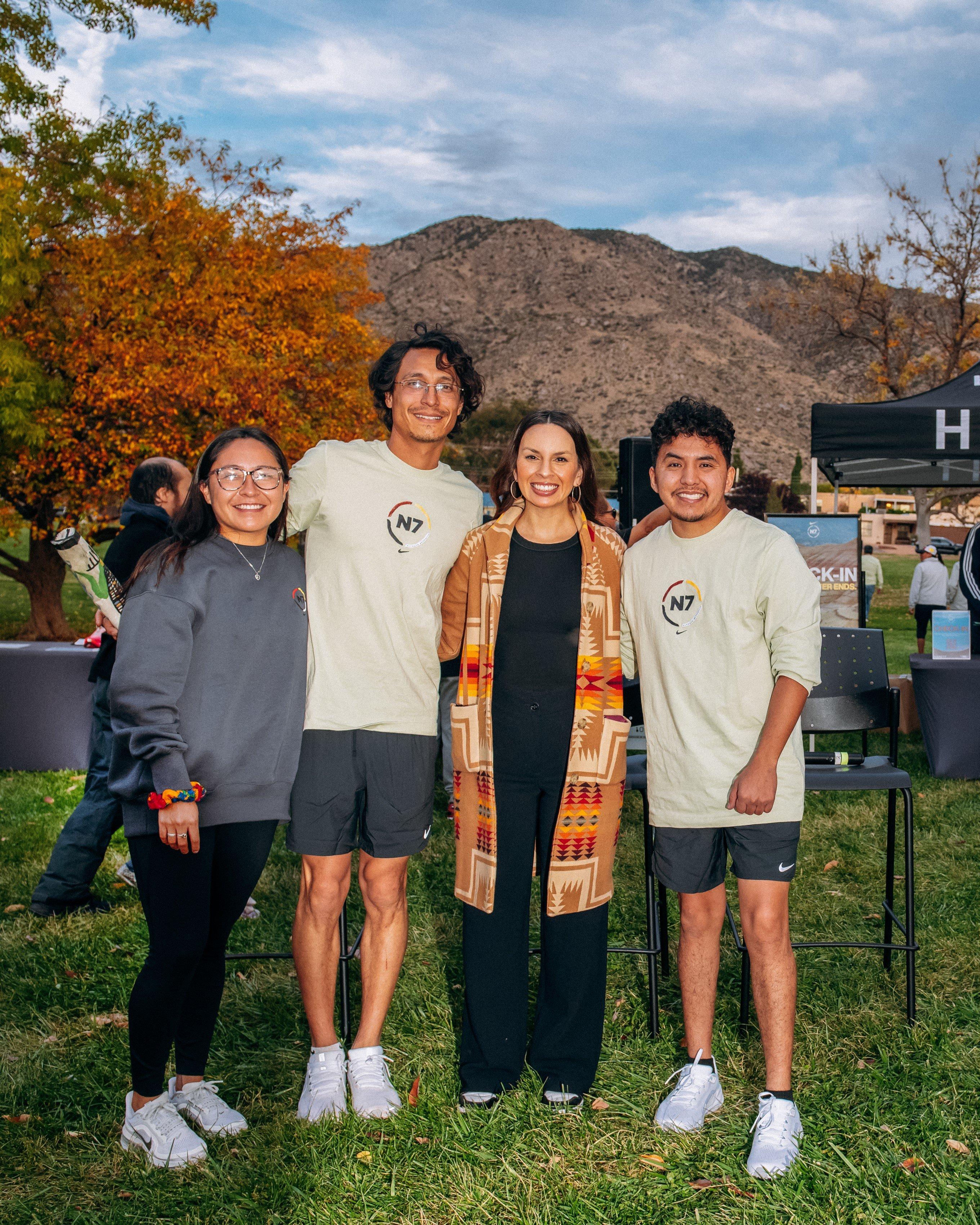 Four people—two young adults on the left and right wearing N7 shirts and two adults in the middle, one wearing a patterned Indigenous shawl—stand smiling in a park with autumn trees and a mountain in the background
