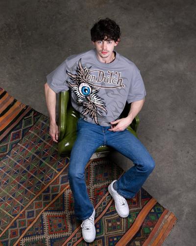 Young man in grey Von Dutch shirt, blue denim pants, and Nike Air Force 1s sitting in a chair.