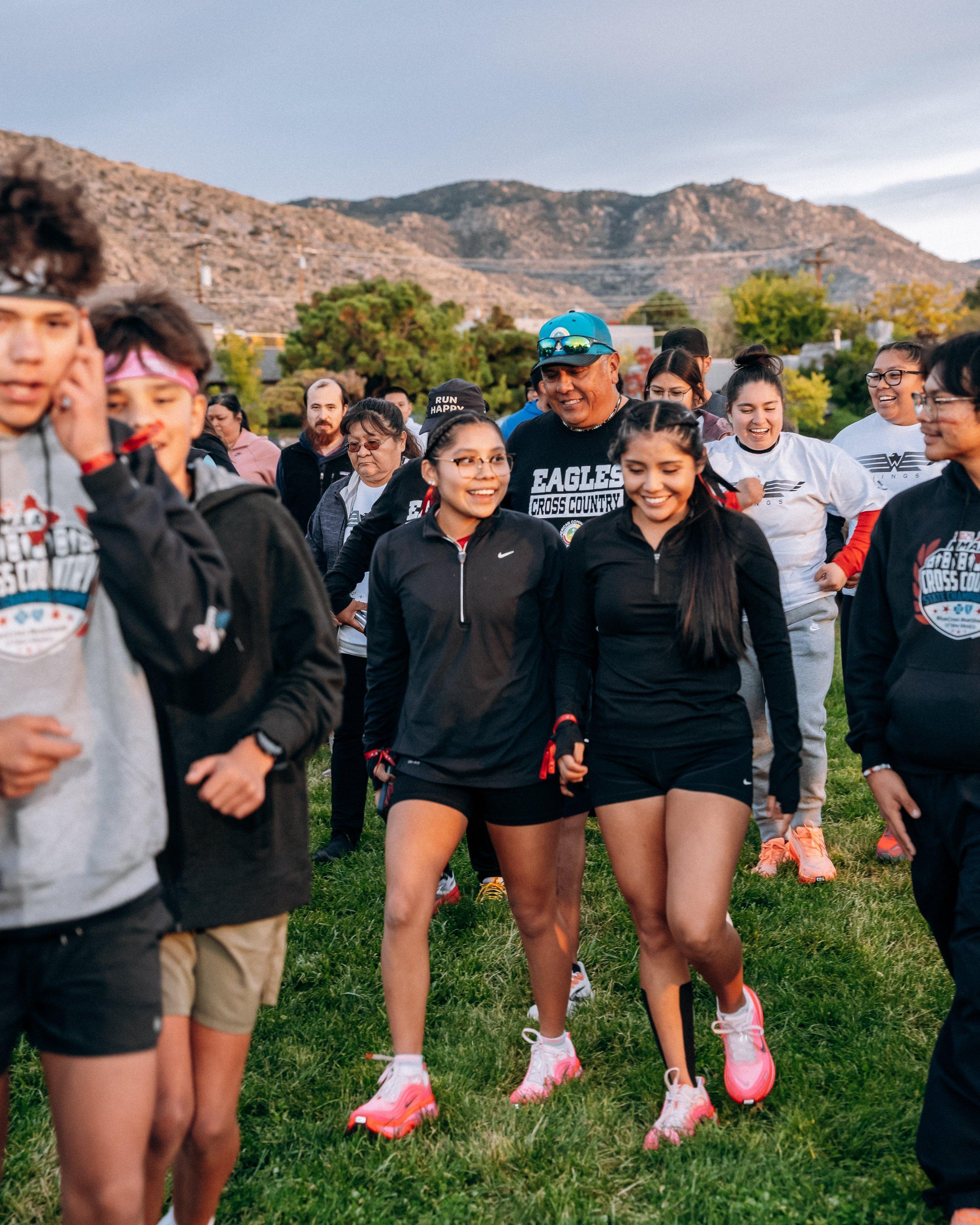 A group of young runners, including high school cross country athletes, walk and talk through a grassy area, smiling as they prepare for the run with mountains visible behind them.