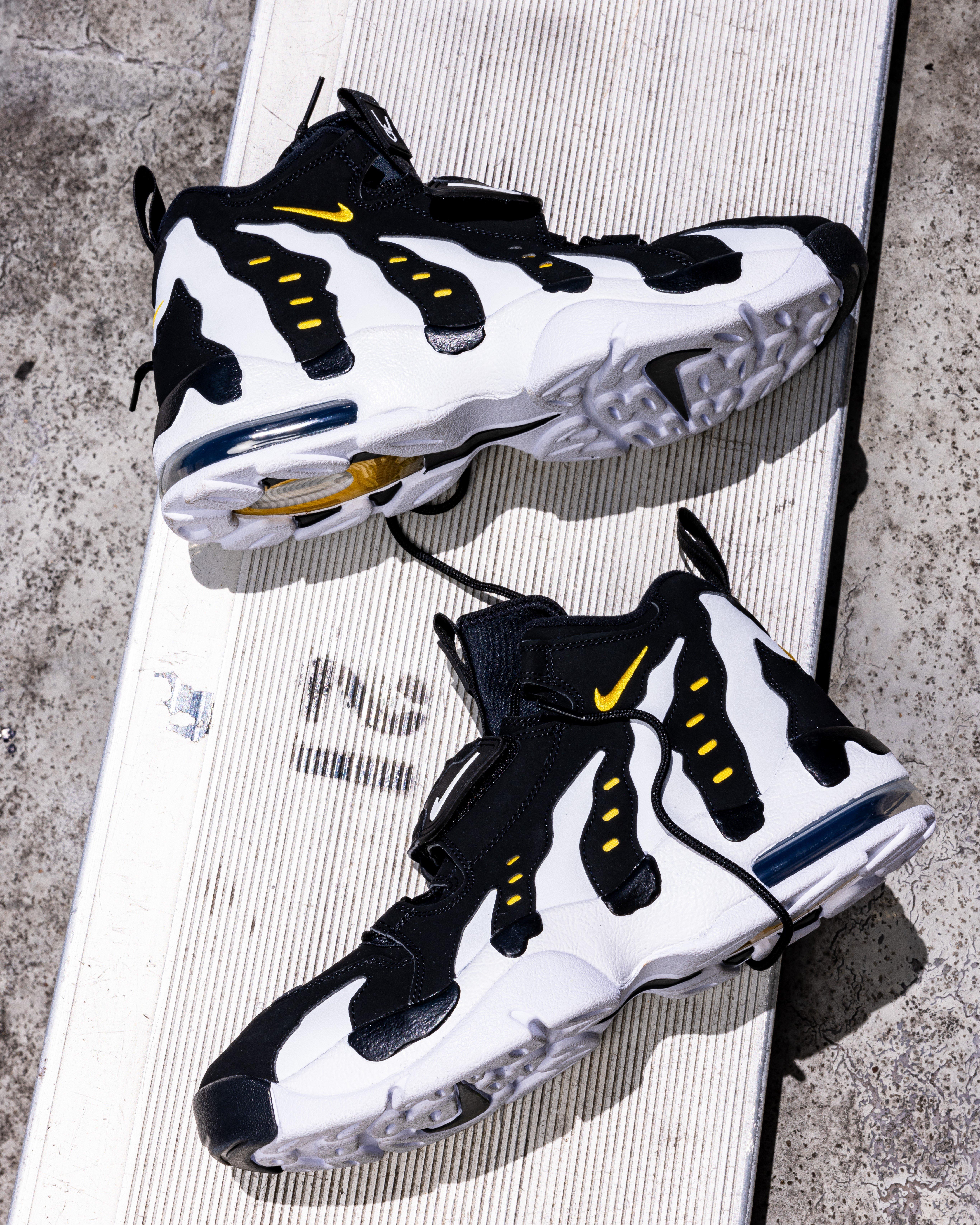 High-angle shot of two Nike Air DT Max '96 sneakers in a black and white "Colorado" colorway, resting on a metallic bleacher. The design highlights bold white claw-like overlays over a black base with small yellow Swoosh accents.