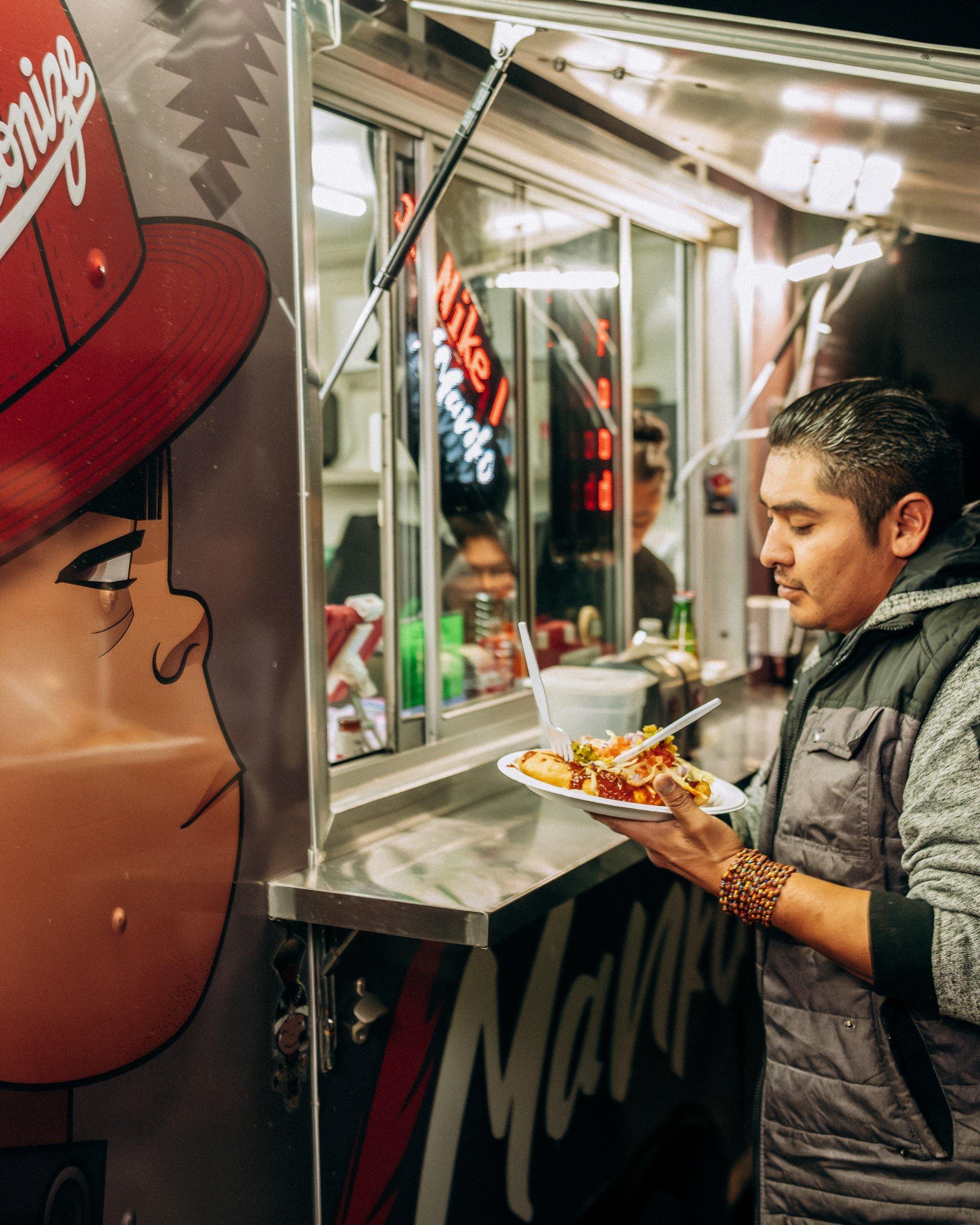 A man holding a plate of food stands at the service window of a food truck. The side of the food truck features a large illustration of a boy wearing a red baseball cap with the word "Decolonize" written on it.