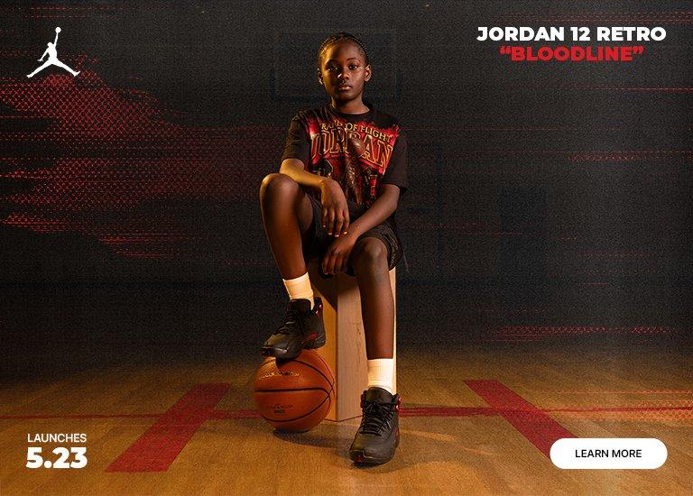 Promotional banner for the kids' Jordan 12 Retro Bloodline launch on 5.23. A child sits on a wooden box with a basketball on a gym floor, alongside a close-up profile shot of the black leather sneakers featuring red metallic accents and white Jordan crew socks.
