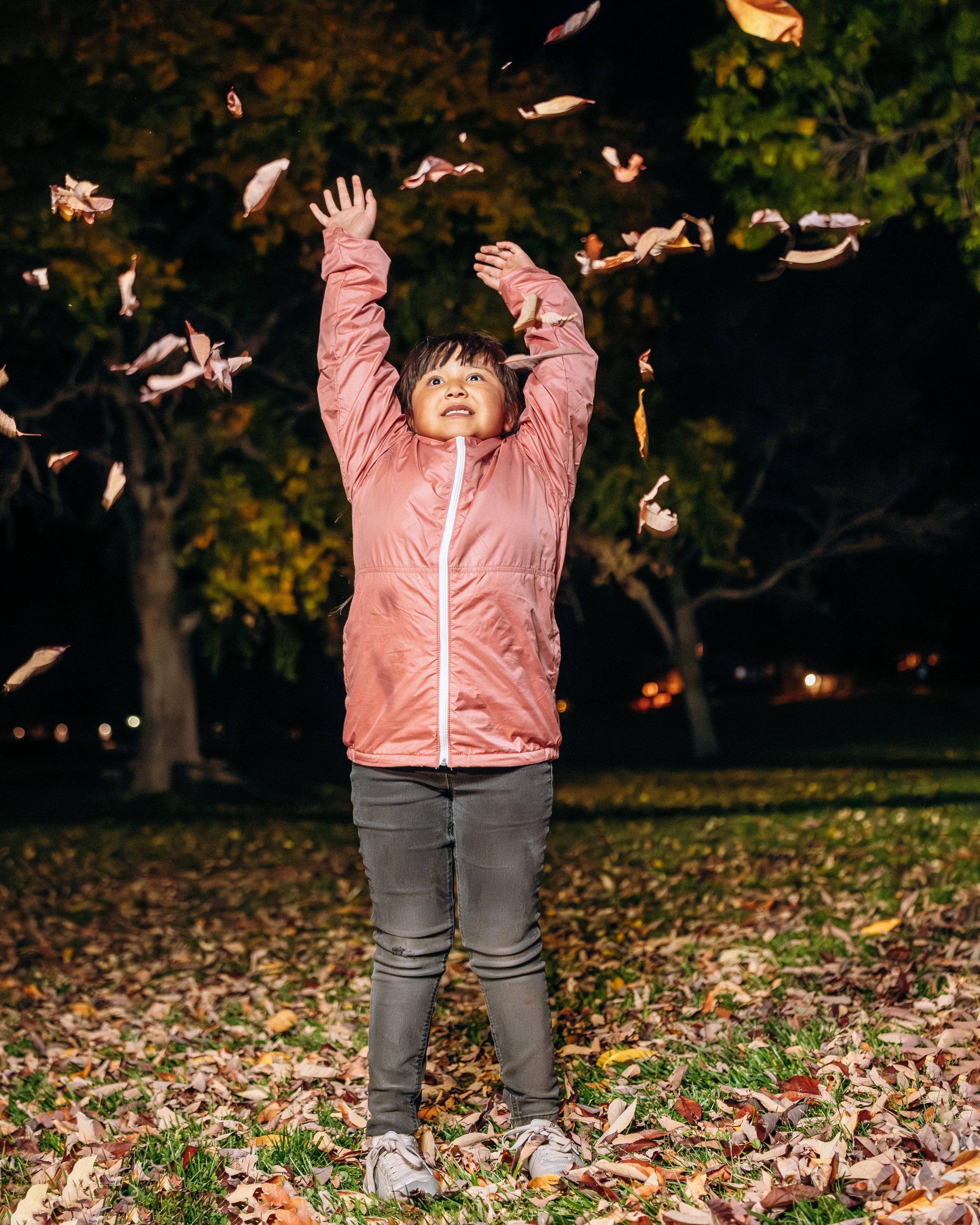 A young child wearing a pink puffer jacket and grey pants stands on a pile of fallen autumn leaves, looking up with arms raised as they throw leaves into the air.