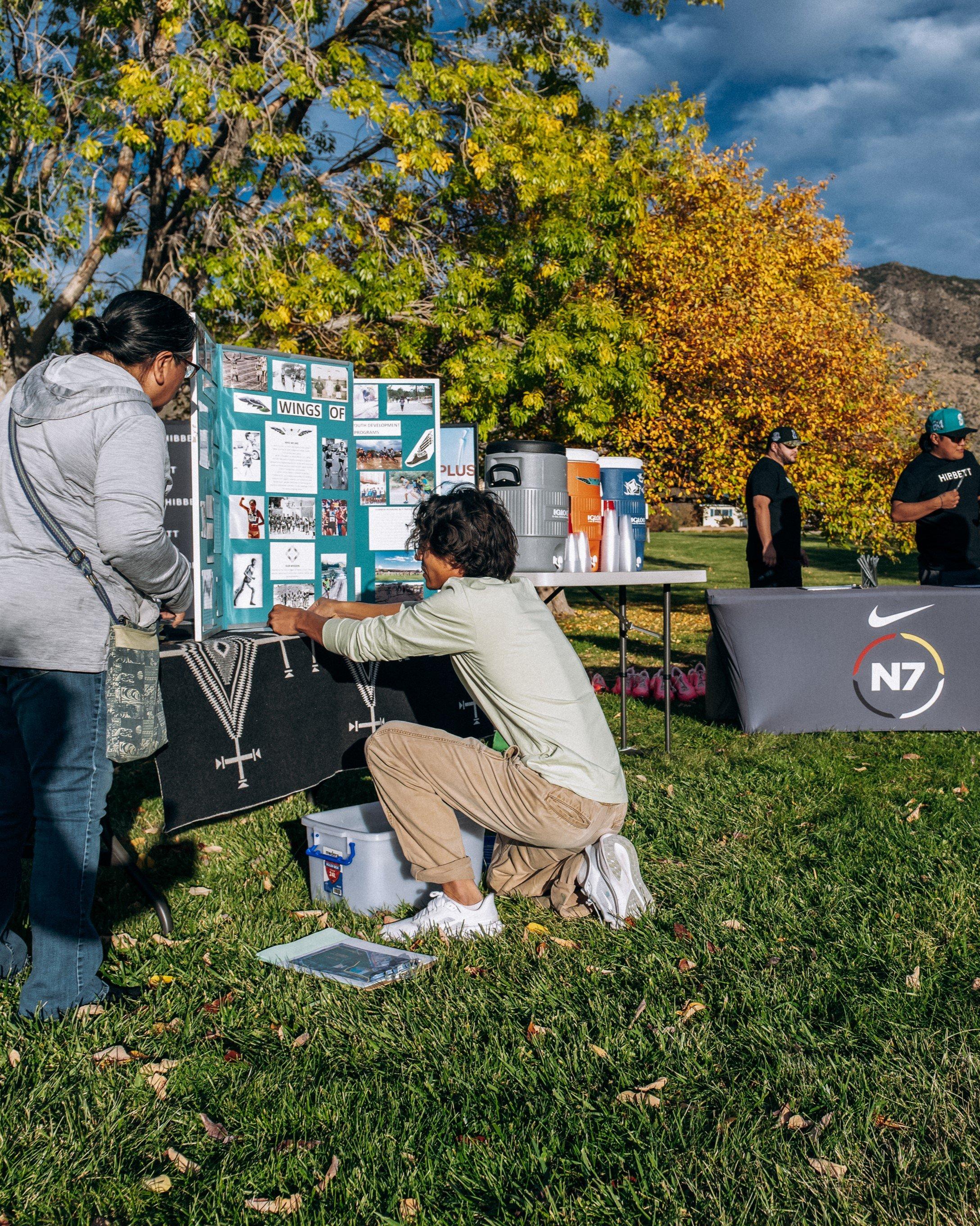Two individuals—a woman standing and a young man crouching—look at a display board set up on a table, which details the work of Wings of America, with event staff and N7 branded tables in the background.