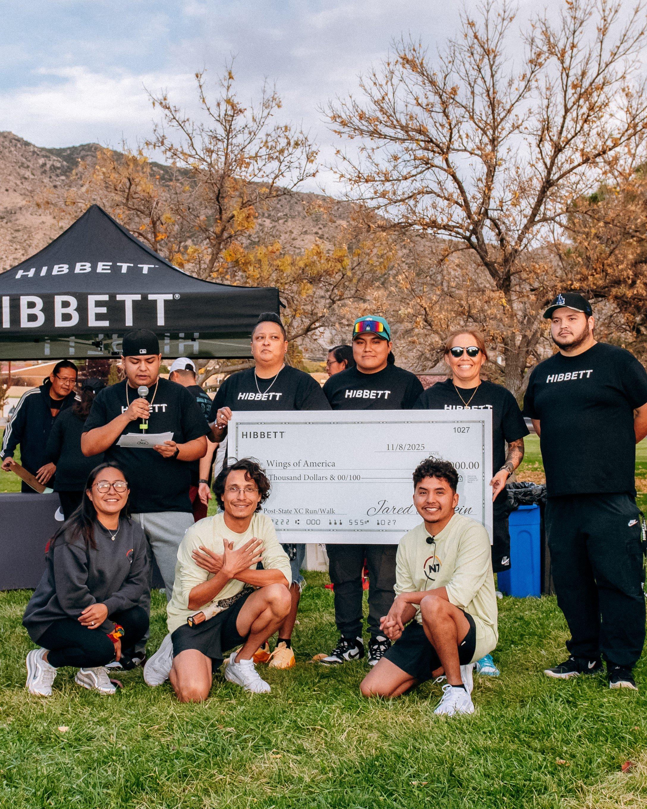 A group of eight people, including some wearing HIBBETT and N7 shirts, stand in front of a HIBBETT tent holding an oversized check representing a $\$15,000$ donation to Wings of America. Mountains and fall foliage are visible in the background.
